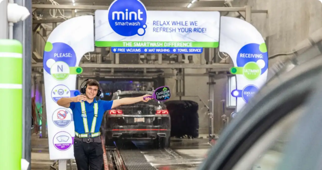Mint Smartwash employee guiding a vehicle through the regular car washes entrance, holding a sign that says 'Continue Forward.